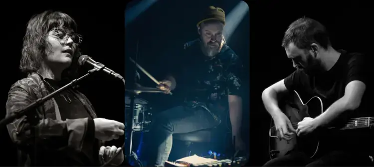 Three musicians are performing: one singing with a microphone, another playing drums, and the third playing an acoustic guitar.