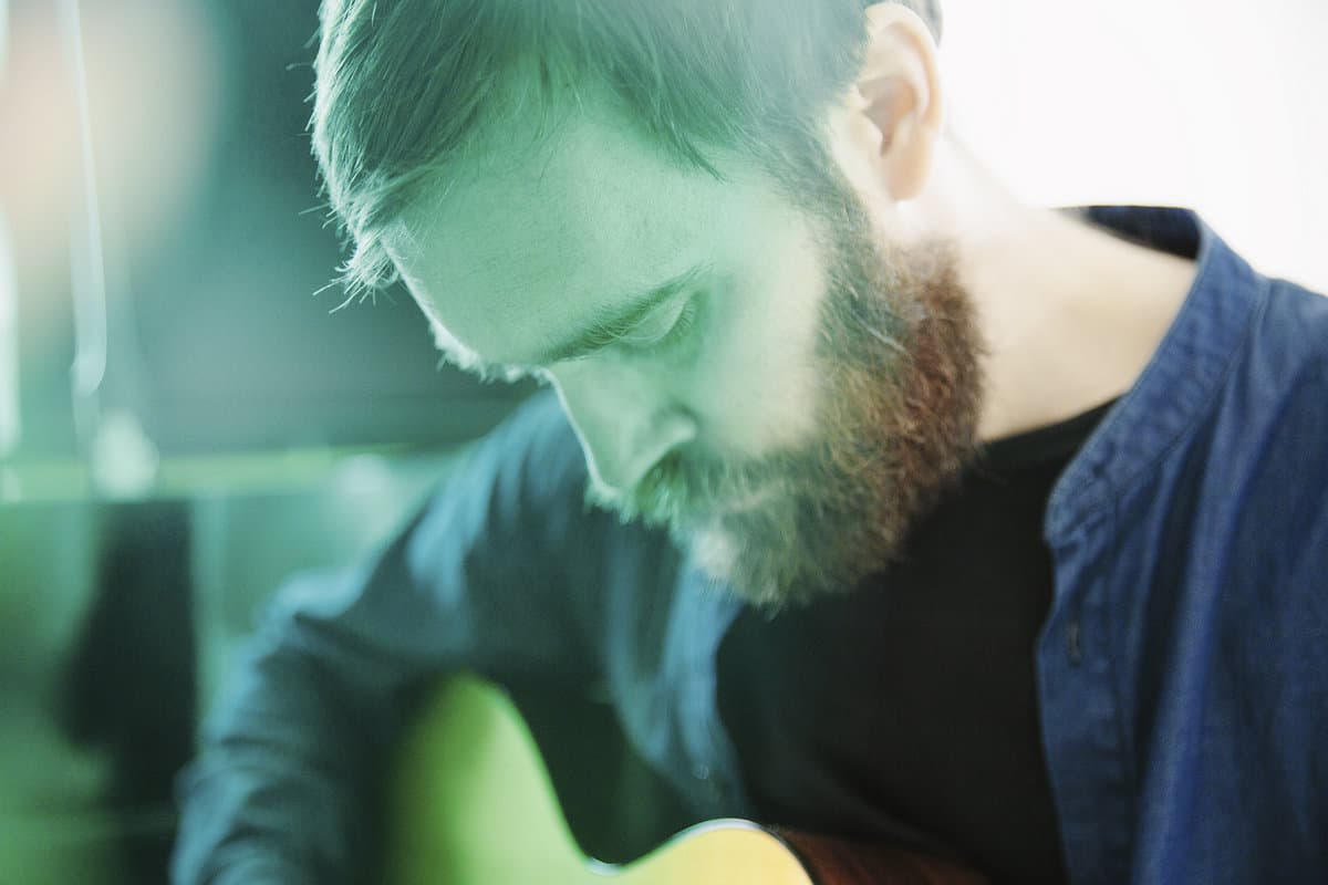 A person with a beard is looking down while playing a guitar, with a greenish tint to the image.